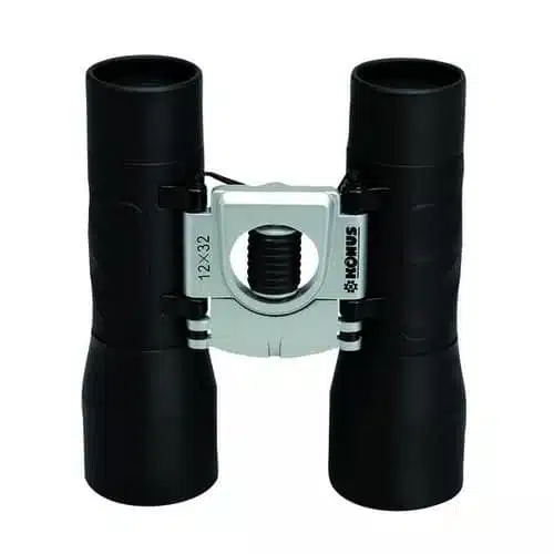 Un par de binoculares "Binocular Basic 2009" negros con un puente plateado que conecta los dos oculares, con la marca "KONUS" y la especificación "12x32". Los binoculares tienen una empuñadura texturizada en cada lado y un botón de enfoque en el centro.