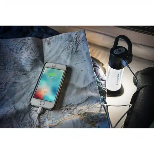 Un teléfono inteligente se encuentra sobre un mapa topográfico, cargándose a través de un cable conectado a una Lámpara Camping Led Lenser Ml6 750lu Bx #500929. La linterna emite una luz suave, iluminando el mapa y el teléfono, que muestra una pantalla de carga. La escena sugiere un entorno al aire libre o de campamento.