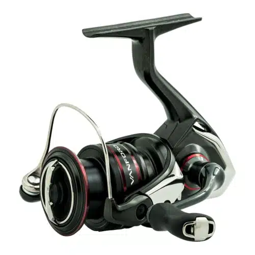 Primer plano de un carrete de pesca moderno y elegante con un acabado negro y metálico. El carrete Shimano Vanford C3000 XG 128m - 0,20mm cuenta con un mango equilibrado, agarre ergonómico y un detalle rojo alrededor de la bobina. El diseño es compacto y aerodinámico, lo que sugiere un alto rendimiento y una artesanía de calidad.
