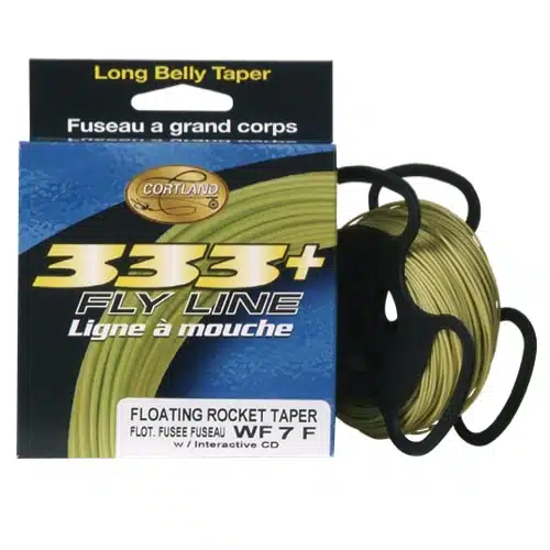 Una caja de línea de mosca Cortland 333+ se encuentra junto a un carrete de línea de pesca verde con la etiqueta "Línea Cortland Fairplay WF7F 27M". El paquete destaca características como "Long Belly Taper", "Floating Rocket Taper" y "WF 7 F" con texto disponible tanto en inglés como en francés. El carrete está asegurado con dos soportes de plástico negros.