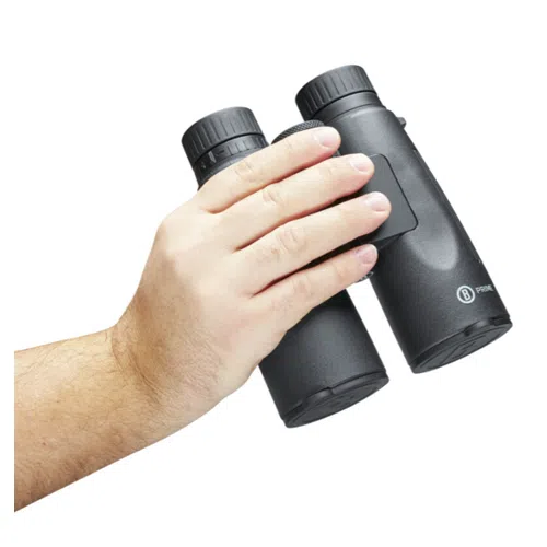Una mano sostiene con confianza el elegante **Binocular Prime 12x50 Bushnell**, mostrando la precisión de su óptica contra un fondo blanco absoluto.