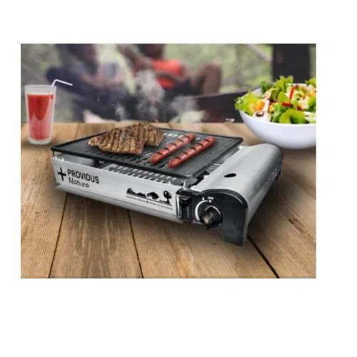 Una Parrilla Camping A Gas Tocoihue Providus adorna la mesa de madera, chisporroteando con salchichas y bistec. Cerca, un bol de ensalada y un vaso de jugo con una pajita se suman al festín, mientras que la vegetación exuberante se asoma por la ventana, completando esta escena gastronómica perfecta.