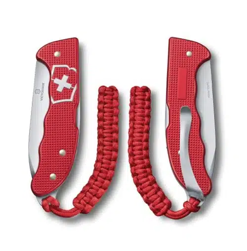 La Cortapluma Hunter Pro Alox Rojo Victorinox 0.9415.20 presenta una hoja plegable y un mango de paracord en un rojo vibrante, que se muestra desde dos ángulos: cerrado y abierto. Este modelo presenta de manera prominente un emblema de cruz blanca en el mango texturizado.