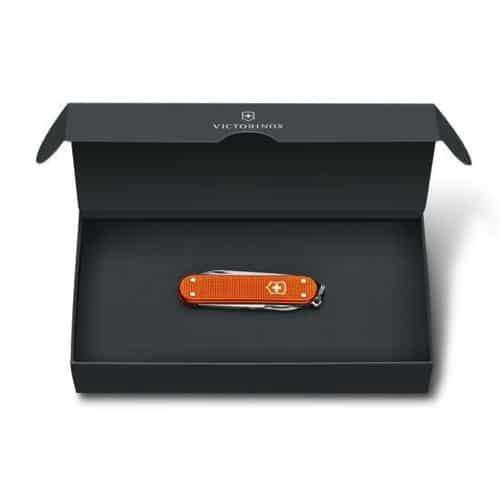 La navaja multiusos Navaja Classic Alox 0.6221.l21, que se presenta en un llamativo color naranja con el logotipo de una cruz blanca, se presenta en una hermosa caja rectangular abierta de color verde oscuro. La tapa superior interior muestra de manera destacada la marca "Victorinox" y el número de modelo 0.6221.l21, lo que realza el encanto atemporal de esta elegante herramienta.