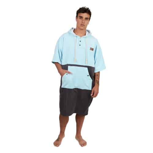 Una persona está de pie, descalza, con el Poncho Terry Kano Celeste, un poncho con capucha de color azul claro y gris, mangas cortas y un gran bolsillo delantero. Tiene una expresión neutra y tatuajes visibles en el brazo izquierdo, que encarnan la sencillez del estilo clásico de Kano. El fondo es blanco liso.