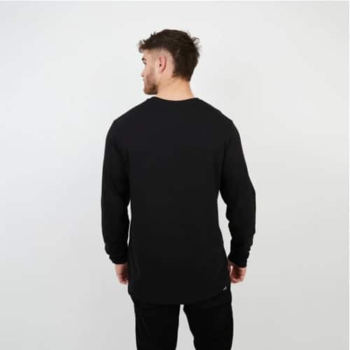Un hombre con cabello corto está de pie, de espaldas, vestido con una elegante Polera Hombre ML Negro Navarino Kano y pantalones negros sobre un fondo sencillo de color claro.