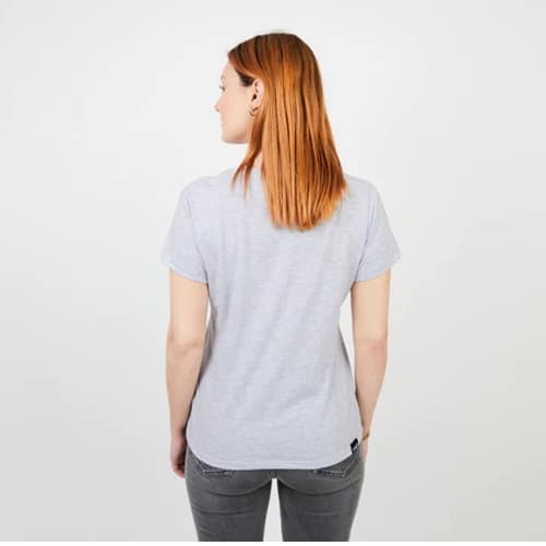 Una persona con cabello rojo está parada de espaldas, vistiendo la Polera Mujer MC Gris Sol Kano y pantalones oscuros, sobre un fondo blanco liso.