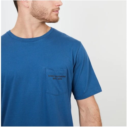 Una persona que viste una camiseta Polera Hombre MC Celeste Tablas Kano tiene un bolsillo en el pecho con el diseño "Kano Outdoors EST. 2003". Tiene la cabeza girada, mostrando una barba corta y un perfil parcial sobre un fondo de color claro.