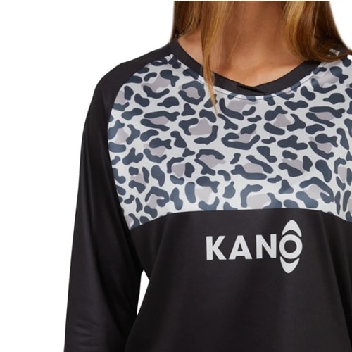 El individuo está vestido con la Polera MTB Farellones W-ML Kano, una camisa negra con un llamativo diseño de estampado de leopardo gris con la palabra "KANO" estampada de forma destacada en el pecho. Posando sobre un fondo blanco liso, la parte superior de su cuerpo irradia un espíritu aventurero similar al de andar en bicicleta por los senderos de montaña de Farellones.
