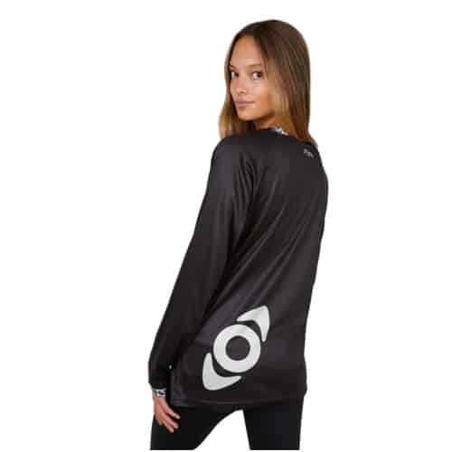 Una persona con el pelo largo lleva la Polera MTB Farellones W-ML Kano, una camiseta negra de manga larga que luce un gran logotipo circular de Kano en la espalda. Está ligeramente girada hacia un lado y mira por encima del hombro contra un fondo blanco inmaculado.