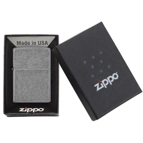 Un encendedor Zippo en una caja negra con el texto "Made in USA" y el logotipo "Zippo" en un lugar destacado. El encendedor Zippo Classic Antique Silver Plate tiene un aspecto de metal cepillado. La tapa de la caja parcialmente abierta muestra con elegancia el envoltorio de este icónico encendedor.