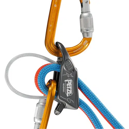 Una vista detallada del equipo de escalada muestra un mosquetón naranja y el Asegurador Petzl Reverso Gris, que asegura cuerdas rojas y azules. El logotipo prominente de Petzl resalta la importancia de las técnicas de escalada seguras con cuerdas bien sujetas.