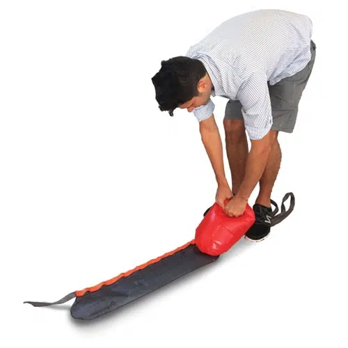Una persona con una camisa a rayas y pantalones cortos está usando una bomba roja para inflar un Parrilla Portaequipaje Inflable Sea To Summit Pack Rack largo y gris que tiene un borde naranja.