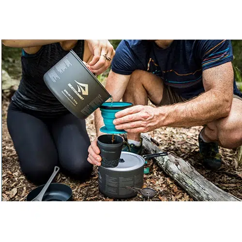 Dos personas están al aire libre preparando una comida con utensilios de cocina para acampar. Una sostiene una olla y vierte con cuidado el líquido en una taza plegable que sostiene la otra. Entre varios utensilios en el suelo, un gotero plegable de silicona Sea To Summit X-Brew Coffee Dripper está listo. Están sentados en un área cubierta de tierra y hojas, inmersos en la serenidad de la naturaleza.