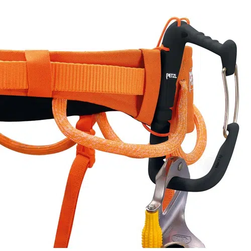 En primer plano, el arnés de escalada Arnés Petzl Hirundos muestra un mosquetón con empuñadura negra acoplado, que resalta sus correas y presillas de color naranja intenso. Un dispositivo de escalada está unido al mosquetón, con el famoso logotipo "Petzl" claramente visible en la empuñadura.