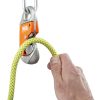 Una mano sujeta una cuerda de escalada amarilla y verde que pasa por un dispositivo de aseguramiento Petzl naranja, que se asegura con un mosquetón Petzl Easytop Steel. Este robusto dispositivo garantiza la seguridad en la escalada en roca.