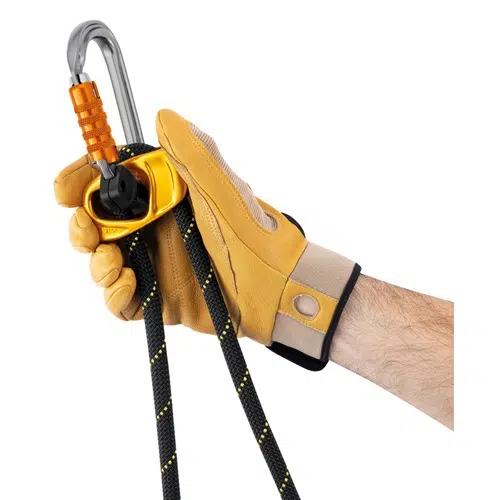 Una persona que lleva un guante amarillo agarra una cuerda de escalada negra y amarilla integrada con un Elemento de Amarre Industrial Petzl Progress Adjust -I, que cuenta con un mosquetón plateado con un seguro naranja y un sistema de poleas amarillo.