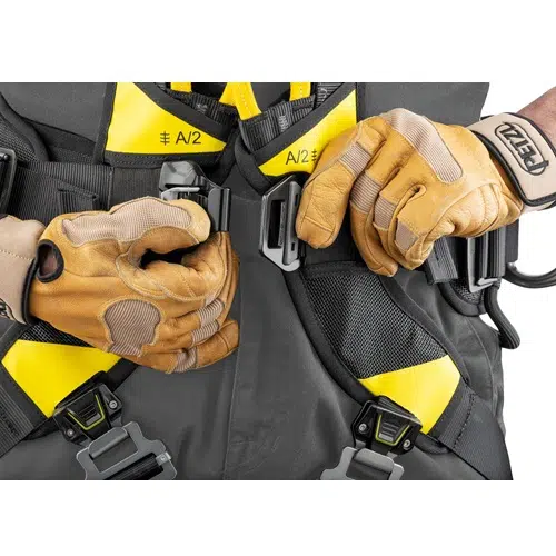Primer plano de unas manos con guantes color canela que ajustan la hebilla de un arnés Arnés Industrial Petzl Volt Europeo. El arnés es oscuro con detalles en amarillo y cuenta con clips adicionales y correas ajustables para un ajuste seguro. La persona lleva equipo de protección.