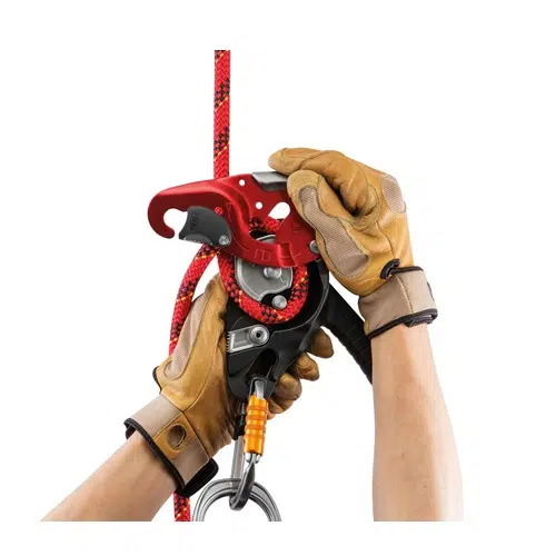 Dos manos enguantadas ajustan un dispositivo de escalada Petzl rojo y negro, posiblemente un Autofrenante IDL o un Descensor Industrial, conectado a una cuerda a juego. El dispositivo parece ser una herramienta de aseguramiento o ascenso. Las manos llevan guantes de seguridad marrones con soportes para las muñecas, lo que garantiza un manejo seguro.