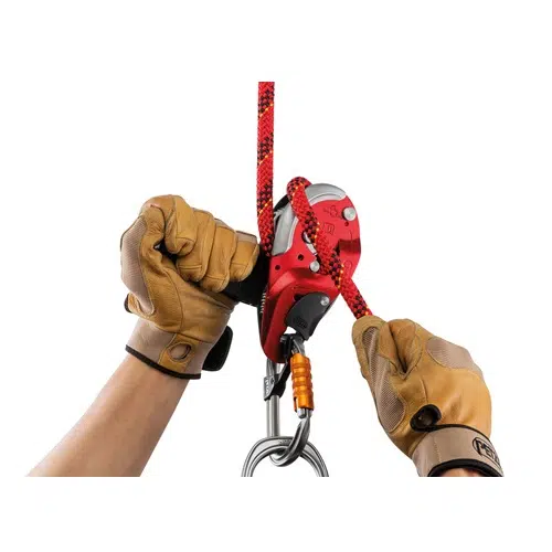 Unas manos con guantes color canela manipulan con destreza un dispositivo de aseguramiento rojo Petzl, una herramienta industrial esencial para descensores, conectado a una cuerda de escalada roja y negra. Un mosquetón naranja está firmemente conectado debajo del dispositivo sobre un fondo blanco.