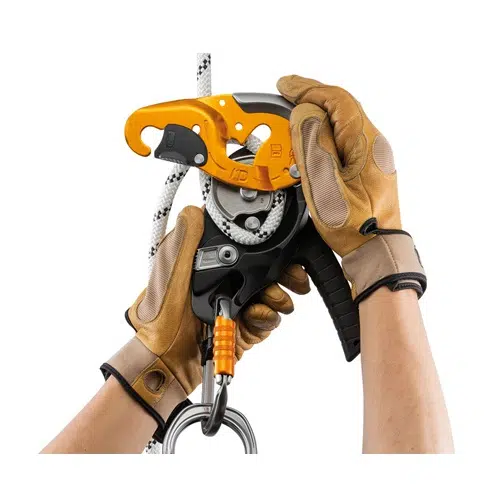 Unas manos con guantes marrones ajustan cuidadosamente un descensor industrial Petzl con una cuerda. El dispositivo cuenta con una palanca naranja y un lazo metálico en la parte inferior, lo que garantiza un manejo seguro durante las escaladas.