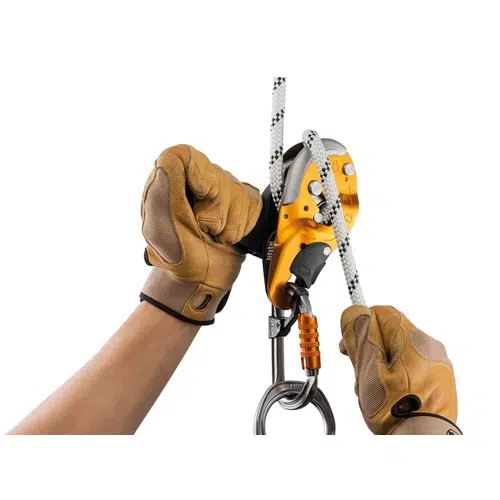 Una persona con guantes marrones muestra una técnica de escalada con un dispositivo de seguridad Petzl amarillo y plateado. La cuerda está colocada con destreza a través del descensor IDS y sus manos están posicionadas para controlarlo.