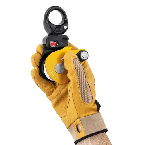 Una mano se pone un guante amarillo y beige mientras sostiene el dispositivo de polea de escalada Polea Industrial Simple Petzl Spin L1, que se distingue por su anilla de sujeción negra. Esta polea industrial, marcada como "Spin L1", está equipada con un exclusivo botón rojo para facilitar su manejo.