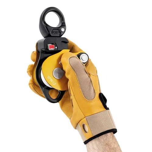 Una persona con un guante amarillo y marrón sostiene un Polea Industrial Simple Petzl Spin L2, un elegante dispositivo mecánico negro y amarillo. Este equipo industrial cuenta con un lazo de metal en la parte superior y un botón rojo visible.