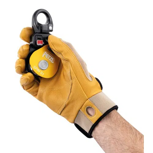 Una mano que lleva un guante amarillo y marrón sujeta una polea industrial Petzl Spin S1 negra y amarilla. El guante, diseñado para uso en exteriores o de seguridad, cuenta con una zona de muñeca reforzada, que complementa la construcción robusta del dispositivo de polea.