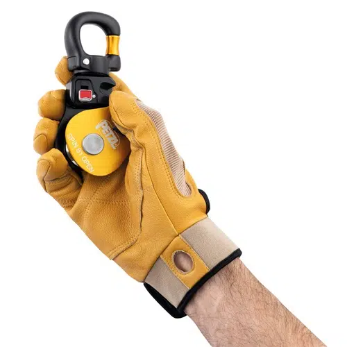 Una mano con un guante de cuero color canela sostiene un dispositivo de seguridad amarillo y negro con un mosquetón, similar al Polea Industrial Simple Petzl Spin S1 Open. El dispositivo tiene un botón redondo plateado y está etiquetado como "SWIVEL OPEN" (abierto giratorio) en texto blanco sobre un fondo blanco.