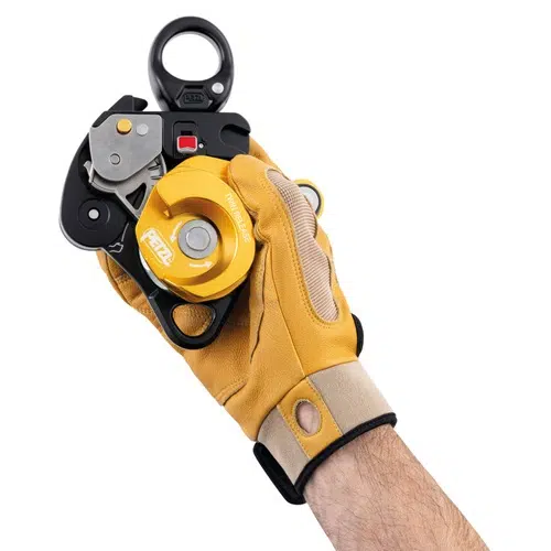 Una persona que lleva un guante de trabajo color canela sujeto con una correa de velcro en la muñeca sujeta la Polea Industrial Doble Petzl Twin Release. Esta polea de rescate negra y amarilla presenta varios componentes metálicos, como una rueda y un ojal para sujetar la cuerda.