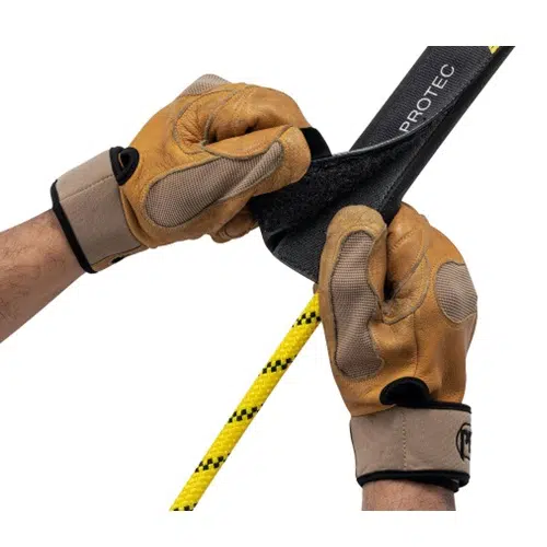 Con gran precisión, unas manos con guantes de protección maniobran con destreza un Protector para Cuerda Fija Petzl Protec negro para asegurar o ajustar una cuerda amarilla y negra. Los guantes color canela con detalles negros complementan perfectamente el dispositivo, garantizando seguridad y un manejo preciso de la cuerda.