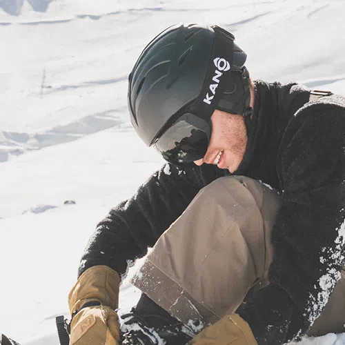 Un esquiador con el elegante casco para nieve HK1 Kano Negro y gafas de esquí se sienta en la nieve, sonriendo mientras se ajusta el equipo. Lleva una chaqueta negra, pantalones beige y guantes marrones. La luz del sol brillante y las montañas cubiertas de nieve crean un telón de fondo impresionante.