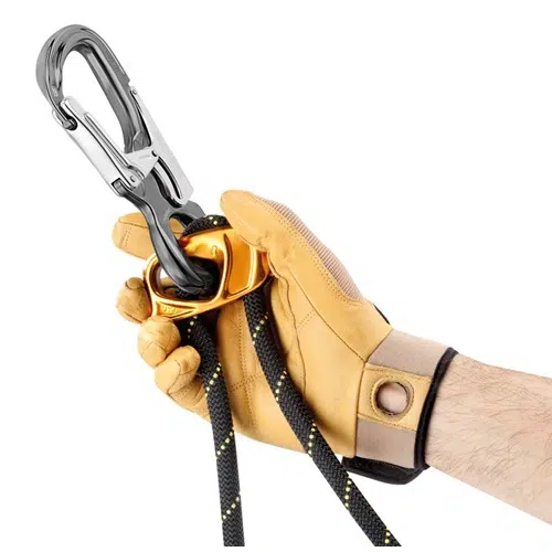 Una mano enguantada sostiene el elegante Conector Petzl Eashook Open, conectado sin esfuerzo a un anillo naranja y una cuerda negra adornada con reflejos amarillos.