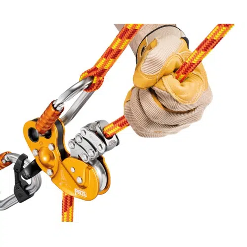 Una mano enguantada sujeta una cuerda semiestática de color naranja y amarillo vibrante, enhebrada a través de un sistema de poleas Petzl plateado y naranja. El equipo incluye mosquetones, lo que sugiere un equipo de escalada o rescate.