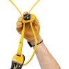 Una mano enguantada sostiene el dispositivo mecánico amarillo y negro conocido como Salvarramas para Poda Petzl Eject, que está sujeto a cuerdas amarillas. Esta herramienta, que lleva la marca "Petzl", probablemente utilizada para escalar o por seguridad, se destaca sobre el fondo blanco liso.