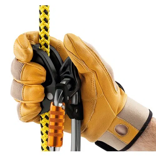 Una mano enguantada sujeta una cuerda de escalada con un patrón amarillo y negro, asegurada con un mosquetón y un dispositivo de seguridad de color naranja. Los guantes son de color tostado con detalles en negro y complementan perfectamente el Elemento de Amarre Industrial Petzl Microflip, lo que lo hace ideal para actividades de escalada o rapel.