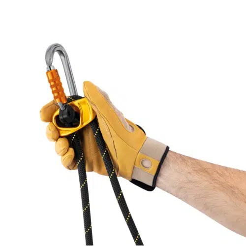 Una mano con un guante amarillo sujeta una cuerda negra que pasa por el dispositivo Cabo de Anclaje Regulable Petzl Progress Adjust-I, que se asegura con un mosquetón plateado. El fondo blanco liso acentúa el colorido del equipo.