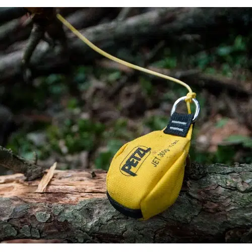Una Hondilla de Lanzamiento Petzl Jet amarilla con base negra está unida a una cuerda fina y descansa sobre la rama de un árbol caído en un bosque. El fondo está difuminado con follaje y ramas verdes.