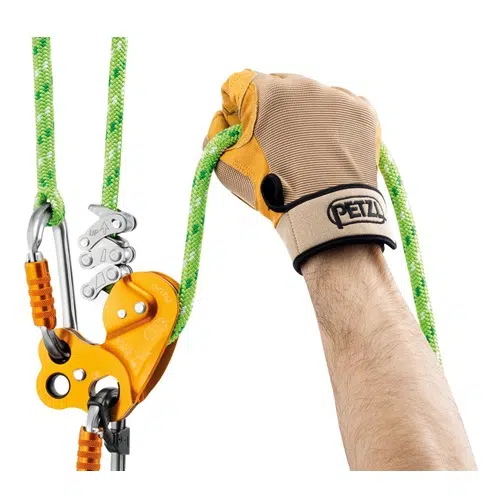 Una mano con un guante de color canela y negro sostiene una cuerda de escalada verde conectada a un Descensor Industrial Petzl Zigzag naranja con mosquetones de metal. Esta configuración se utiliza para escalar o para realizar tareas de seguridad con arnés.