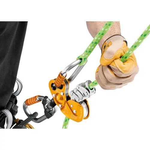 Una persona con guantes beige maniobra hábilmente a través de un sistema de cuerdas para escalar, utilizando dispositivos mecánicos de color naranja y plateado, entre los que se encuentra el Descensor Industrial Petzl Zigzag Plus. El conjunto incluye mosquetones y se conecta a un arnés, lo que lo convierte en un equipo esencial para la escalada industrial o las actividades de rescate.