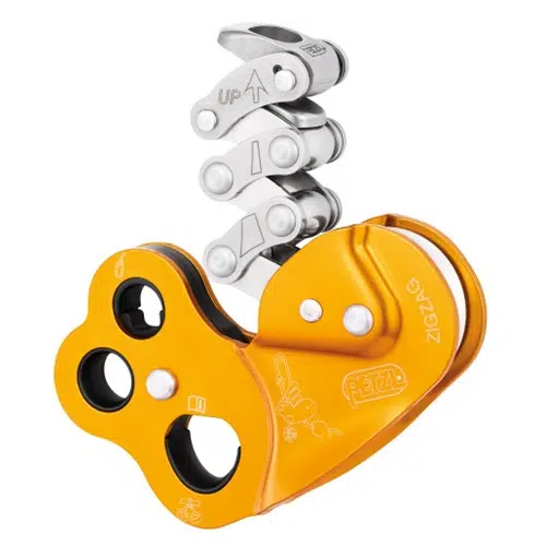 Primer plano de un dispositivo mecánico de color naranja brillante con componentes negros y plateados. Parece ser una herramienta de montaje industrial con múltiples ranuras y piezas móviles, marcada con la marca "Descensor Industrial Petzl Zigzag".