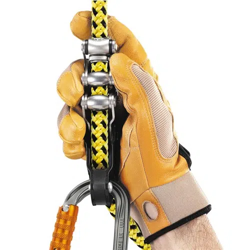 Una mano que lleva un guante de color canela y beige sujeta firmemente una cuerda trenzada amarilla y negra que pasa por un sistema de poleas de metal, con un mosquetón naranja adjunto. Esta configuración se asemeja al Elemento de Amarre Industrial Petzl Zillon, ideal para escaladas precisas o operaciones de rescate.