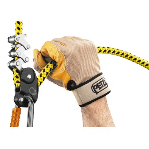 Una mano enguantada agarra una cuerda en una instalación de escalada o de seguridad, con el Elemento de Amarre Industrial Petzl Zillon. El guante de color canela brinda protección y muestra la marca "Petzl" en la muñeca. La cuerda amarilla, resaltada con patrones negros, pasa a través de un dispositivo metálico equipado con múltiples poleas.