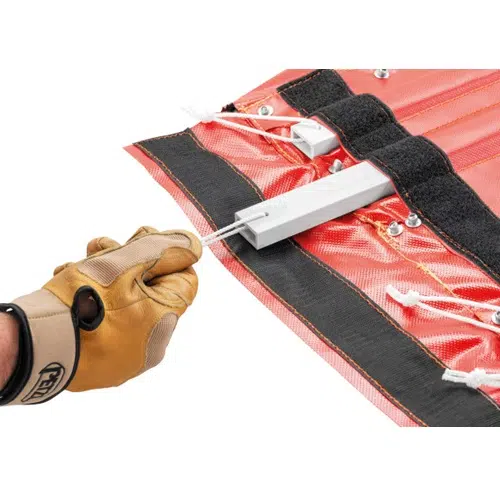 Una persona con un guante de cuero marrón tira de una cuerda atada a la Camilla para Rescates Petzl Nest. La tela roja y negra presenta ojales metálicos y cordones blancos, con una pieza metálica rectangular asegurada en ella, lo que la hace ideal para situaciones de emergencia.
