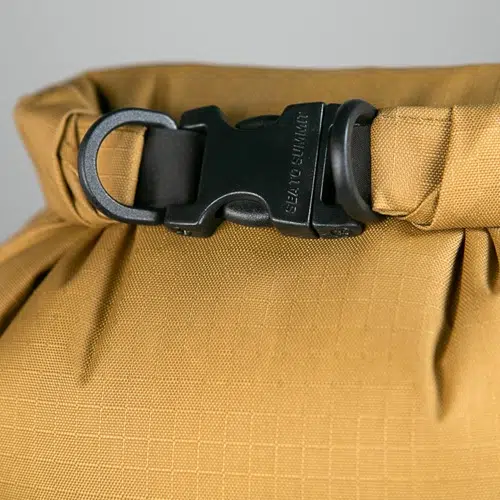 Primer plano de una bolsa impermeable Big River de color marrón amarillento de Bolsa Sea To Summit con una hebilla y correa de plástico negras. La hebilla con acabado mate tiene un texto grabado parcialmente visible, mientras que la tela presenta un patrón de cuadrícula sutil.