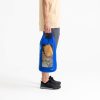 Una persona sostiene una bolsa impermeable ligera Sea To Summit de color azul, que tiene una ventana transparente que muestra los artículos en su interior, incluido un trozo de tela amarilla. Está vestida con una camisa beige, pantalones cortos grises y zapatillas negras sobre un fondo blanco liso.
