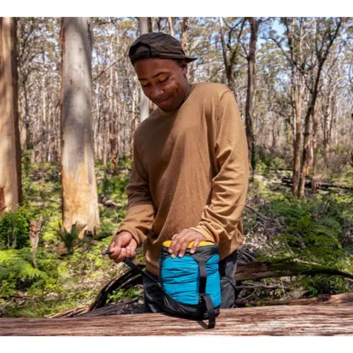 Una persona con una camisa marrón de manga larga y una gorra negra sostiene una Bolsa de Compresión Sea To Summit Evac Compression Dry Bag azul con una correa para el hombro en un entorno forestal. Está rodeada de árboles altos y una vegetación exuberante y parece estar preparándose para una aventura al aire libre.