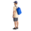 Una persona, vestida con una camiseta beige, pantalones cortos grises, zapatos negros y una gorra azul marino, se encuentra de lado, mostrando su mochila azul vibrante con la parte superior enrollada. Su estilo y funcionalidad recuerdan a la Bolsa Estanca Ligera Sea To Summit. El fondo blanco realza la simplicidad de la escena.