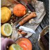 Un cuchillo Opinel N°10 VRN de acero carbono con mango de madera yace abierto sobre un papel arrugado, rodeado de limones, una mandarina, almendras y nueces. En primer plano, una mano sostiene un vaso de jugo de naranja recién exprimido.